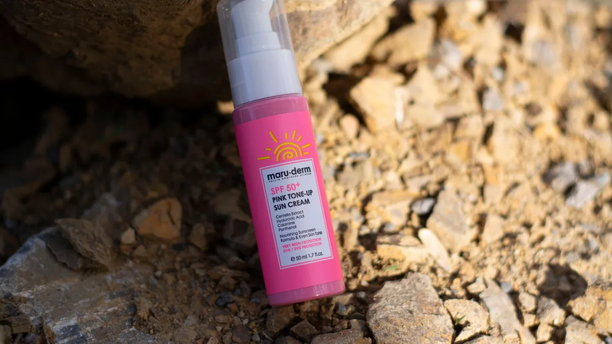 Close-up of SPF 50+ sun cream bottle with pink toned packaging on rocky surface.