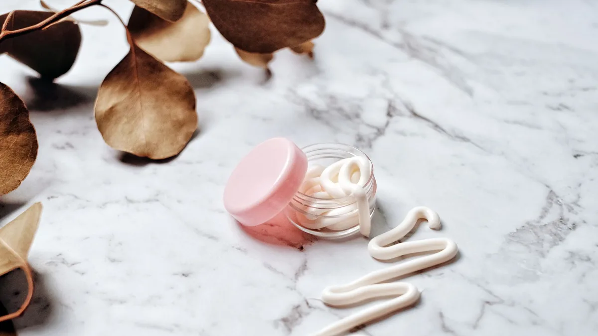 Close-up of skincare cream in a pink glass jar on a marble surface surrounded by leaves, ideal for beauty and wellness themes.
