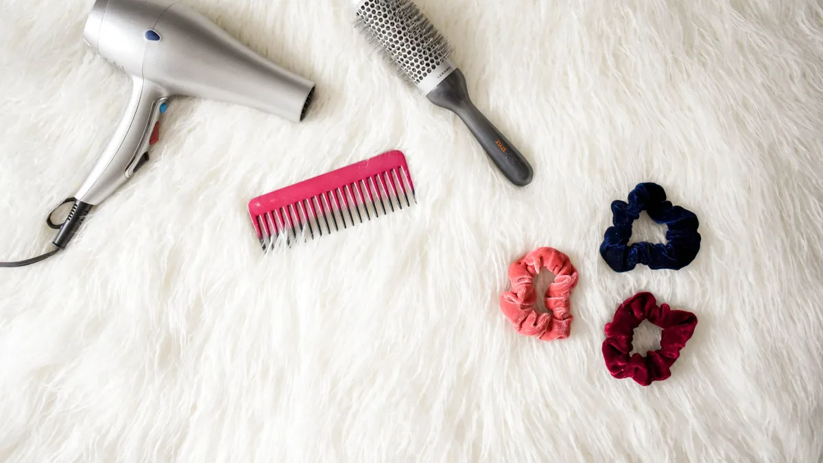 Flatlay of hairstyling tools including a hairdryer, brush, comb, and scrunchies on white fur.