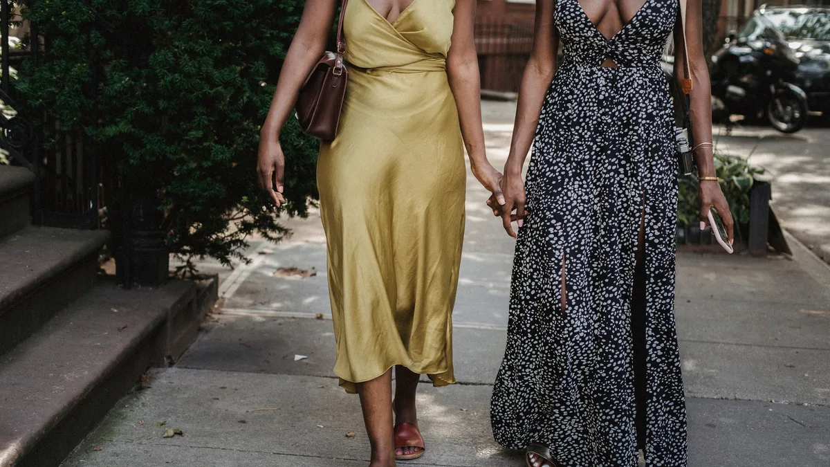 Two fashionable women in maxi dresses holding hands while walking through an urban sidewalk.