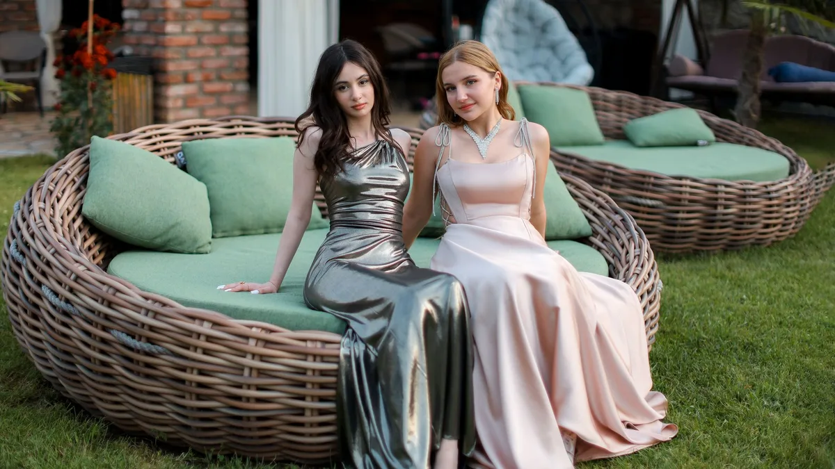 Two women in glamorous evening dresses relax on stylish outdoor furniture, exuding elegance and poise.